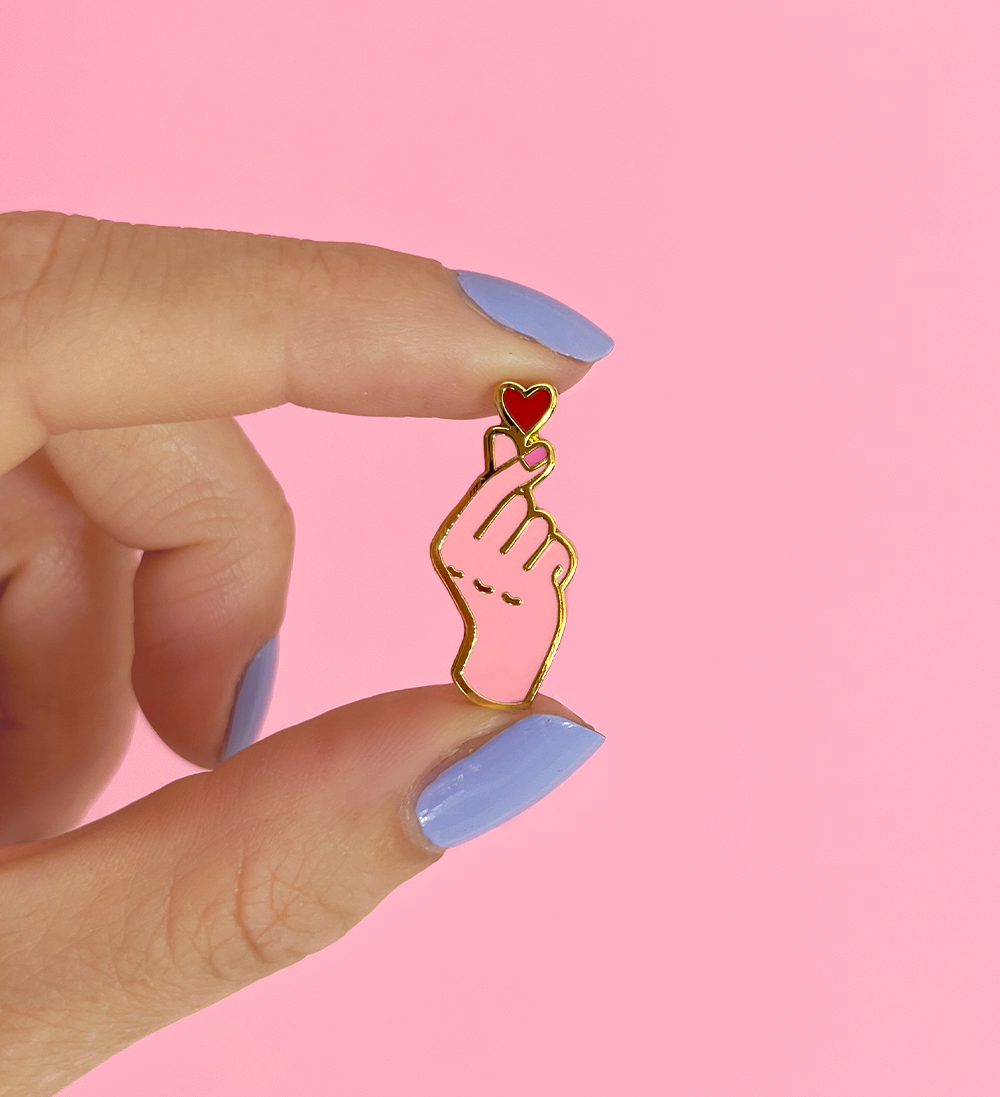 Pins, main coeur avec les doigt, signe K-pop. Pins en émail doré symbolisant l'amour avec le symbole coréen, 2 doigts qui forme un coeur. Un petit coeur en sort. Les couleurs sont rose. Le pins est en mal dur, la couleur est lisse et le pins résistant.